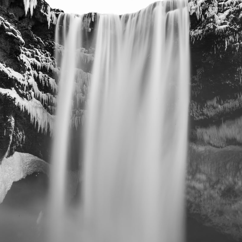 Black and white image of the top of a waterfall from below. Icicles are hanging from the edges of many rock ledges around and behind the waterfall.